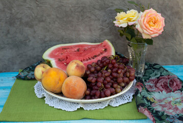 On a platter is a half of a watermelon, grapes and peaches; nearby in a crystal vase are yellow roses..