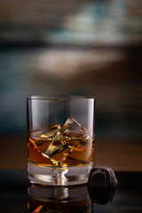 A glass of Kentucky Bourbon on rocks and piece of chocolate candy  placed on black reflection surface with colored wood background
