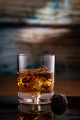 A glass of Kentucky Bourbon on rocks and piece of chocolate candy  placed on black reflection surface with colored wood background