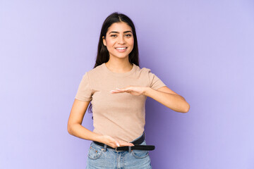Young indian woman isolated on purple background holding something with both hands, product presentation.