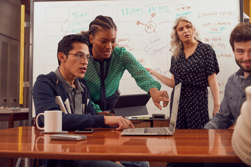 Young female leading group of young co-workers in discussion of brand strategy, in conference room with digital tablets and laptop  in front of dry erase board with flowchart and calendar ..