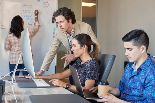 Co-Workers talking at computer in office while woman is working on dry erase board with flowchart in background 