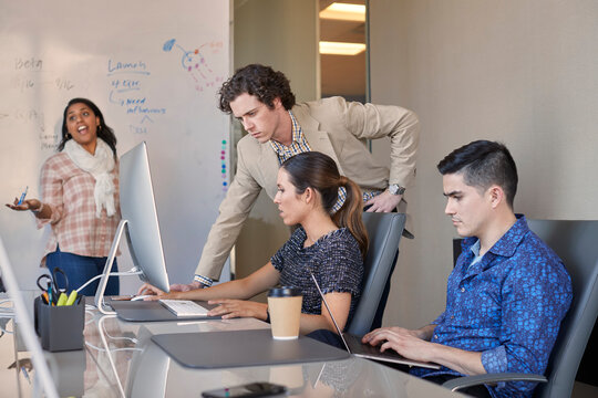 Co-Workers talking at computer in office while woman is working on dry erase board with flowchart in background 