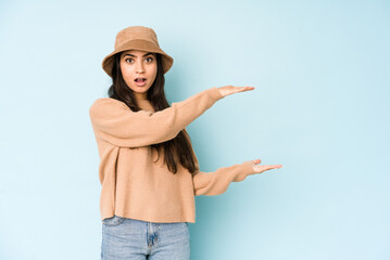 Young indian woman wearing a hat isolated on blue background shocked and amazed holding a copy space between hands.