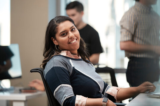 Young woman sitting at computer in office looking to the camera smiling, co-workers having discussion around dry erase board in background 