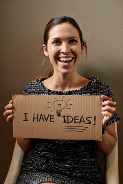 Portrait of Young woman sitting in chair holding cardboard sign the reads I have Ideas, looking off camera 