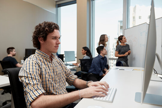 Young man using computer in office looking, co-workers having discussion in background 