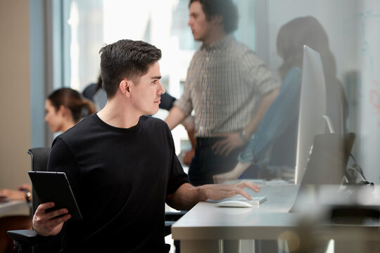 Young man multi-tasking by sitting at computer in office and checking tablet, co-workers having discussion around dry erase board in background 