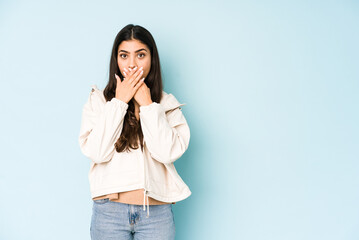 Young indian woman on blue background shocked covering mouth with hands.