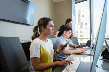 Young woman using computer in office typing on keyboard, and checking mobile phone with co-workers working in background 