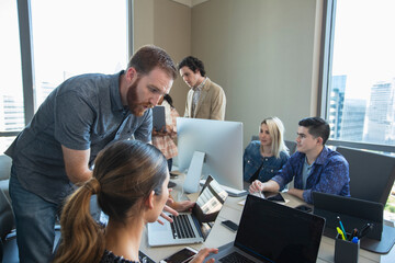 worker in office conference room providing tech support to colleague working on laptop, co-workers working in background 
