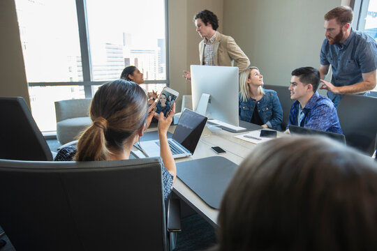 Woman taking selfie of herself and co-worker with mobile phone, while colleagues work in background of conference room 