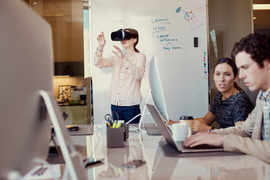 Co-Workers working on computer in office while woman is using Virtual Reality Headset standing in front of dry erase board with flowchart in background 