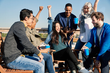 Group of young co-workers hanging out on rooftop patio having a drink and celebrating achievement reached on cell phone 