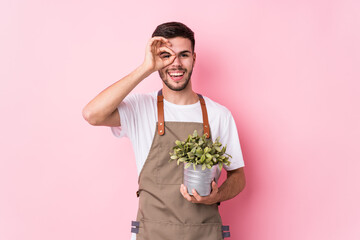 Young caucasian gardener man holding a plant isolated excited keeping ok gesture on eye.