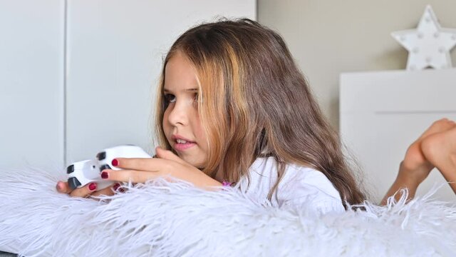 A little girl is playing a computer console on a sofa in a bright room. Concurrent games for children. Joystick for control in hands. Emotions and fun of the child. Copy space.High quality 4k footage.