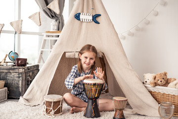 Little drummer girl playing on djembe sitting in front of ethnic tent at home