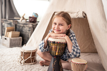 Cute little girl with ethnic drums sitting near wigwam in playroom