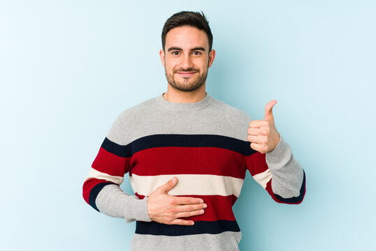 Young caucasian man isolated on blue background touches tummy, smiles gently, eating and satisfaction concept.
