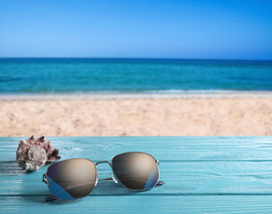 Fototapeta na wymiar Shell and stylish sunglasses on turquoise wooden table near sea with sandy beach, space for text