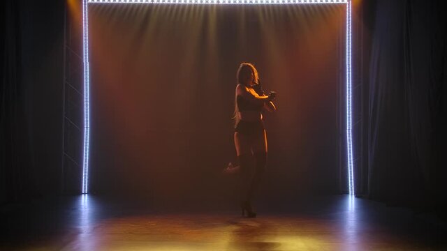 Passionate young Caucasian brunette dancing captivatingly in black womanish lingerie in a dark studio with staged light. Silhouette of a slender toned body, slow motion.