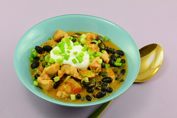 Black bean chicken soup with dollop of sour cream in blue bowl with golden spoon on lavender background