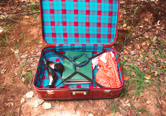 Open vintage suitcase with women's retro dress, high-heeled shoes and books, lying on the ground in the summer forest