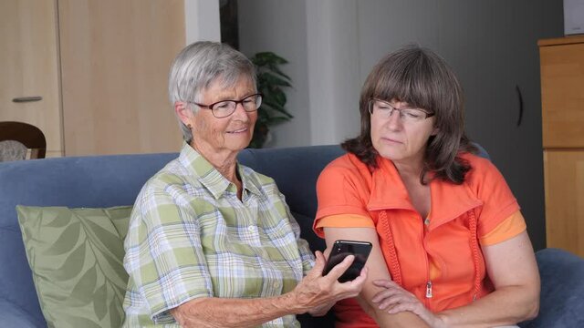 Grandmother practices using smartphone with her adult daughter on a sofa in the living room