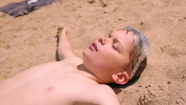 A little boy sunbathing by lying carefree on the sand next to the lake shore.