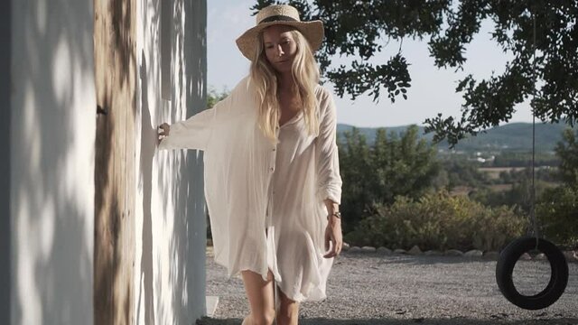 Young woman with long dress and straw hat walking barefoot alongside an old wall on a rural farm in Ibiza, Spain. Closing in from the front in slow motion.