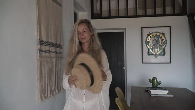 Young woman with long dress walking and spinning straw hat in a Bali style house. Leading shot in slow motion.