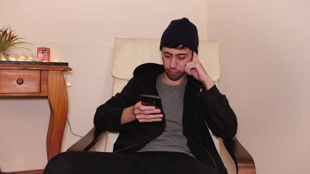 Bored young man with short beard and a dark beanie using his smartphone