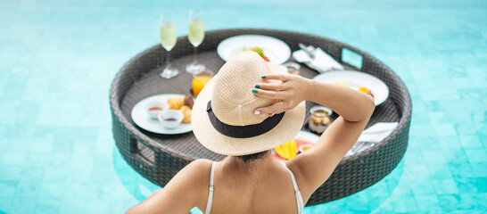 Happy woman in white swimsuit having Floating Breakfast tray in luxury pool hotel, young female with hat enjoy in tropical resort. Relaxing, Exotic summer travel, holiday, vacation and weekend