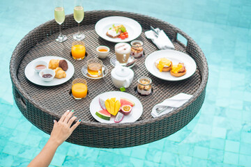 Floating Breakfast tray in swimming pool at luxury hotel or tropical resort villa, fruits; mango, watermelon dragon and passion fruit, food, bread, coffee, tea, wine and orange juice. Exotic summer