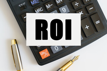On the table there is a pen, a calculator and a business card on which the text is written ROI. RETURN ON INVESTMENT