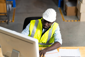 middle aged African American warehouse worker working on personal computer and preparing a shipment in large warehouse distribution centre