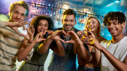 Fun night. Young men and women showing heart signs while posing together for camera. Multiracial group of friends hanging out at party in the bar