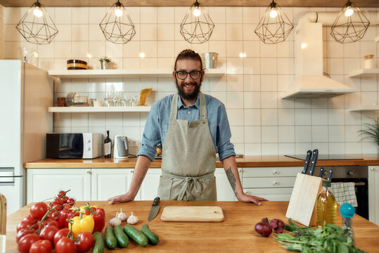 Young Man, Italian Cook In Apron Smiling At Camera While Preparing Healthy Meal With Vegetables In The Kitchen. Cooking At Home Concept