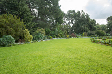 Fototapeta na wymiar View of a large formal garden landscaping with well-tended neat lawn and a variety of trees, flower beds. Background texture of grass and plants in a park. Copy space for text.