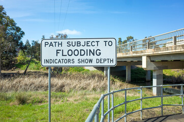 The sign says 'path subject to flooding indicators show depth' on a footpath leads to a bridge underpass. Melbourne, VIC Australia.