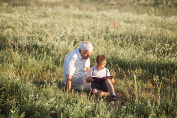 Fototapeta na wymiar Mid shot of grandfather and his grandson while reading a book together in meadow Small boy making first steps in read.