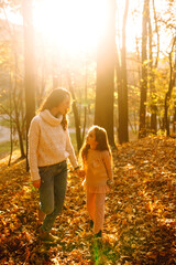 Fototapeta premium Stylish young Mother and daughter walking in the autumn forest at sunset. Mother and daughter embrace in autumn park. Mom and child having fun together in autumn. Family on a walk. 