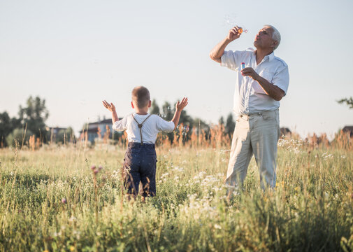 Happy Senior Man Grandfather With Cute Little Boy Grandson Playing In Field.  Male Blowing Soap Bubbles While An Excited Kid Enjoys The Bubbles. Happy Teenage