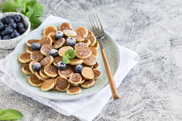 Tiny pancakes for breakfast. Cereal pancakes with blueberries in plate on grey background. Trendy food. Copy space for text or design