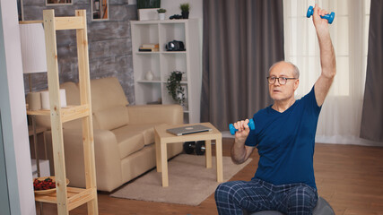 Senior man doing shoulder press with dumbbells in living room. Old person pensioner healthy...