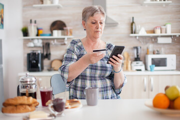 Retired old woman using credit card for online shopping in kitchen during breakfast. Senior elderly...