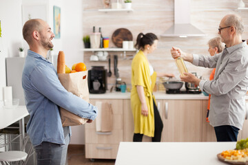 Obraz premium Cheerful man with groceries bag from supermarket in home kitchen. Senior man holding bottle of wine.