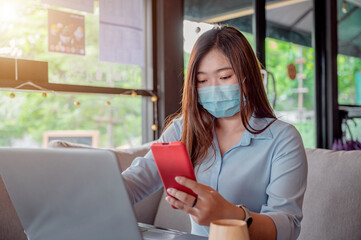 Thai female office worker Put on a mask I am using a red smartphone and working on my laptop in a coffee shop during the day. She was wearing a blue shirt and sipping coffee.