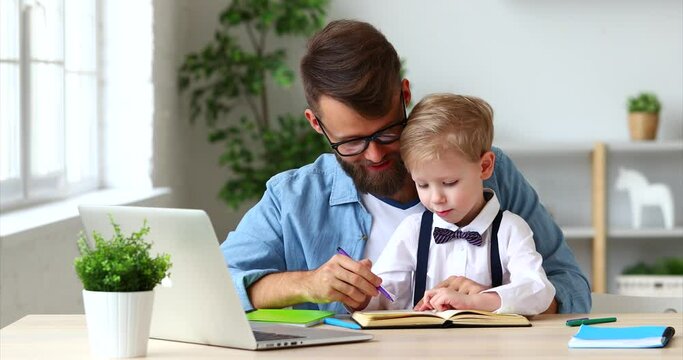 Young bearded father in eyeglasses pteaches a child son to write and hold a pen correctly  during online studying together at home
