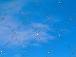 Abstract view of stainless steel mesh fence with clamps and clear blue sky background with light soft white clouds. flat on view. summer scene. modern construction materials concept. fine wire cable.
